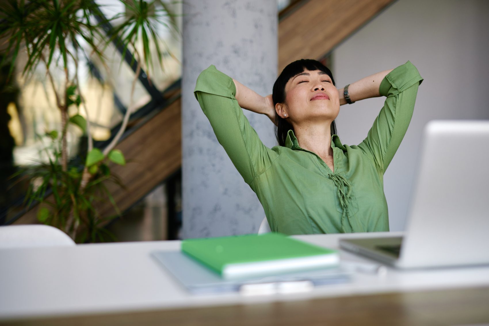 A woman leaning back from her desk, eyes closed and hands behind her head, taking a moment